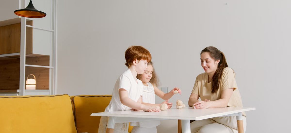 A mother and two children playing blocks on a white table in a cozy living room.
